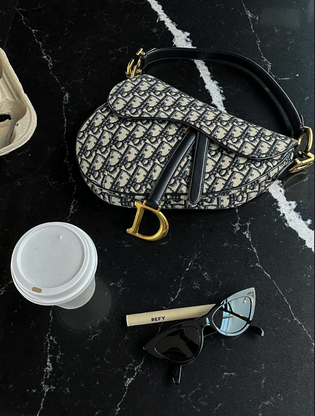  Dior saddle bag oblique monogram gold hardware on a black marble table with black cat eye sunglasses a cup of coffee and refy brow product